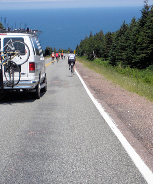 Cyclists riding down a steep hill on Cabot Trail toward the ocean followed by a silver SUV.