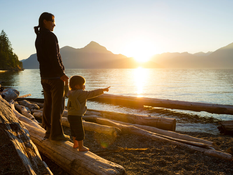 mother and daughter stand on log on beach at sunset