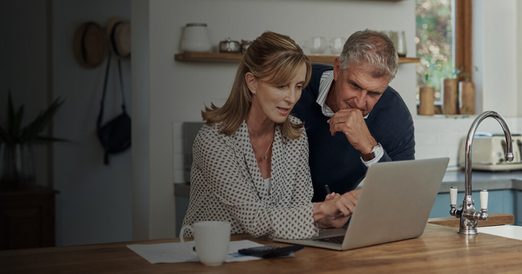 An older couple looking at a laptop with papers beside them on a kitchen counter.